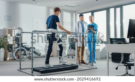 Modern Hospital Physical Therapy: Patient with Injury Walks on Treadmill Wearing Advanced Robotic Exoskeleton. Physiotherapy Rehabilitation Scientists, Engineers, Doctors use Tablet Computer to Help Royalty-Free Stock Photo #2029416692
