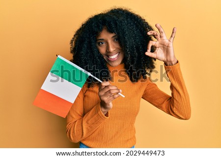 African american woman with afro hair holding ireland flag doing ok sign with fingers, smiling friendly gesturing excellent symbol 