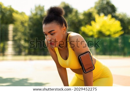 Active Training. Tired African American young woman in yellow sportswear bra and arm band with cell resting during morning run, catching breath standing outdoors at public park or sports field Royalty-Free Stock Photo #2029452275