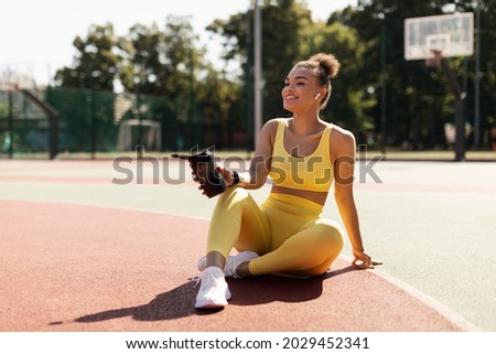Energy, Vitality, Wellness Concept. Smiling African American woman sitting on basketball field ground, holding shaker, drinking water, taking break after outdoors exercise, wearing wireless earbuds Royalty-Free Stock Photo #2029452341