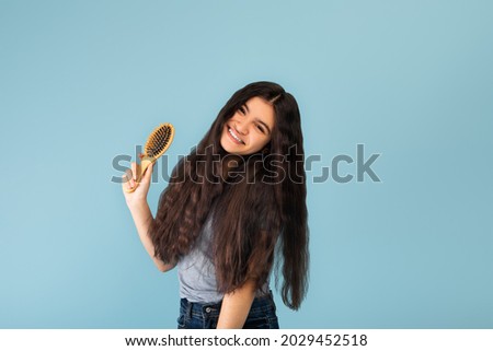 Attractive Indian teenage girl with long silky dark hair holding wooden brush, posing over blue studio background. Female teenager enjoying her beautiful healthy locks