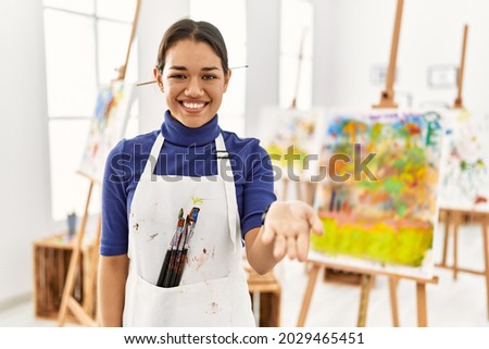 Young brunette woman at art studio smiling cheerful offering palm hand giving assistance and acceptance. 