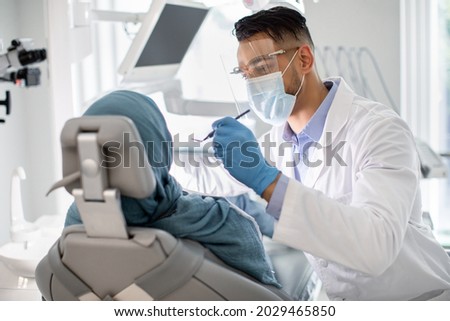 Middle Eastern Male Dentist In Medical Mask And Face Shield Checking Patient's Teeth, Professional Stomatologist Doctor Making Dental Treatment To Islamic Lady In Hijab At Modern Clinic, Closeup Royalty-Free Stock Photo #2029465850