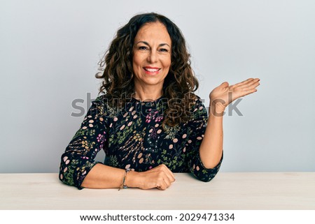 Beautiful middle age woman wearing casual clothes sitting on the table smiling cheerful presenting and pointing with palm of hand looking at the camera. 