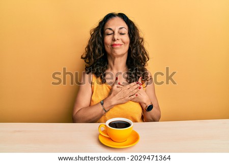 Beautiful middle age woman drinking a cup of coffee smiling with hands on chest with closed eyes and grateful gesture on face. health concept. 