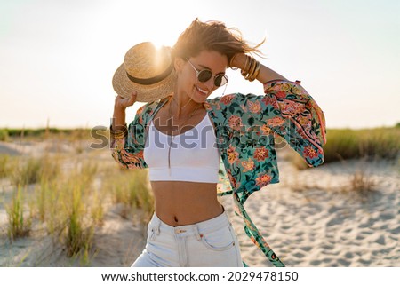 pretty attractive slim smiling woman on sunny beach in summer style fashion trend outfit happy, freedom, wearing white top, jeans and colorful printed tunic boho style chic and straw hat Royalty-Free Stock Photo #2029478150