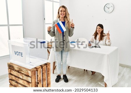 Young blonde woman at political stand holding russia flag surprised with an idea or question pointing finger with happy face, number one 