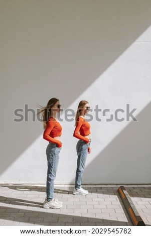 Twin sisters in identical clothes posing against the backdrop of a light wall with contrasting light in urban area