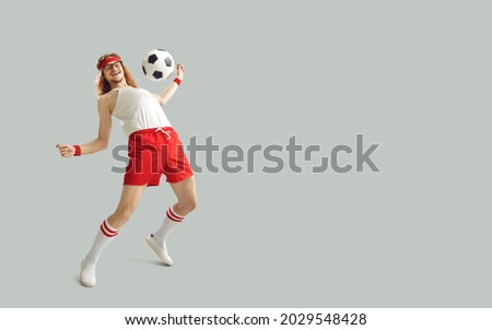 Excited nerdy male sports fan having fun with a football. Happy funny young man in white and red activewear kicking away soccer ball on light gray background with empty space for advertising text