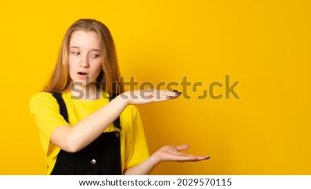 Surprised teen girl shapes height of object, has appealing look, over yellow background. Size and measure concept. Banner copy space.