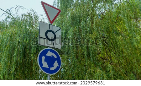 Bent road sign Roundabout, give way near the highway against the background of green tree.