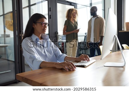 Young mixed race woman, female office worker working on computer while sitting at her workplace in the modern office, colleagues standing on the background