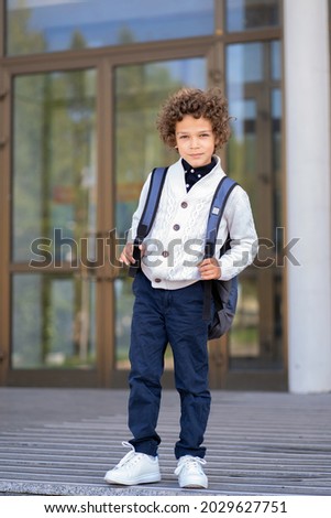 a schoolboy with curly hair stands with a schoolbag next to the school Royalty-Free Stock Photo #2029627751