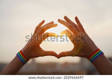 Gay pride, love and marriage concept. Hands with gay pride LGBT rainbow flag wristband making heart sign at sunset sky
