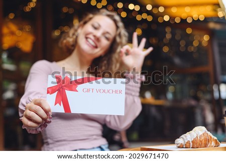 Young happy woman in casual clothes sit at cafe table eat breakfast show ok gesture hold gift certificate coupon voucher card for store relaxing in restaurant during free time indoors. Focus on card Royalty-Free Stock Photo #2029671470