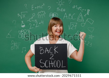 Teacher mature elderly lady woman 55 wear white shirt hold in hand demonstrate sign board poster back to school demonstrating strength power isolated on green wall chalk blackboard background studio