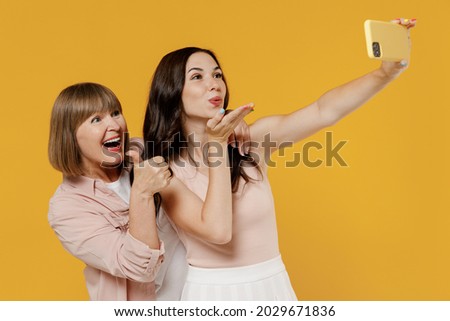 Two side view young daughter mother together couple women wear casual clothes do selfie shot on mobile cell phone blow air kiss show thumb up like gesture isolated on plain yellow background studio