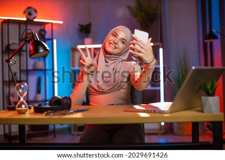 Happy arabian woman in headscarf gesturing sign peace while taking selfie modern smartphone. Young lady sitting at desk with wireless laptop during evening time at home. Remote work concept.