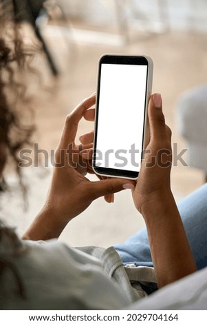 Young African American woman hands holding cell phone with mockup white blank display, empty screen for app ads at home. Mobile applications technology concept, over shoulder close up view.