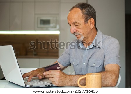 Trendy mature man is working from home with laptop sitting at the table in his kitchen, buying something with credit card online, happy retirement, home-office concepts
