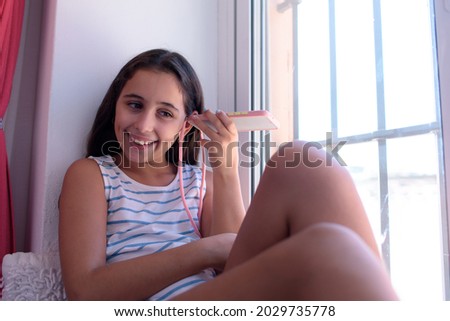 Pretty brunette girl, sitting by the window of her house, smiles as she listens to a voice message, bringing the speaker of her smartphone close to her ear.