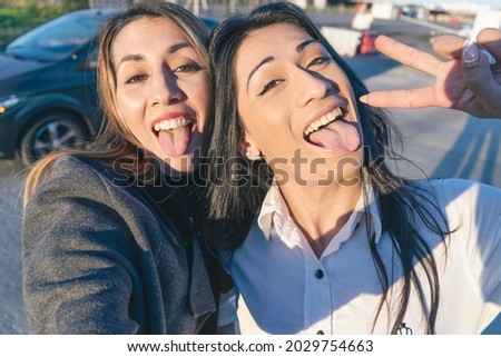 Two beautiful Latina women friends sticking out their tongues and taking a selfie with their cell phone. friendship and camaraderie concept.