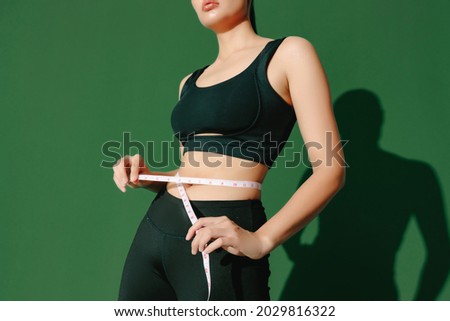Crop close up body of sporty woman measuring her thin waist with a tape measure isolated on green background. Dynamic movement. Strength and motivation. Royalty-Free Stock Photo #2029816322