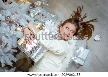 an attractive young adult woman, blonde, in a white dress, is holding a gift in a silver package. A woman is filmed in close-up near a Christmas tree. The concept of the holiday.