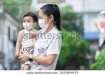 Asian mother wearing white face mask holding her little toddler baby girl who is her daughter wear baby face mask, they looking out, COVID-19 concept