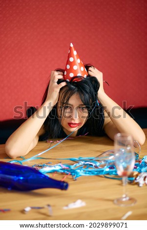 Young woman with red rubbed lipstick and cap, sitting tired at table in messy room after birthday party, woman after party at home