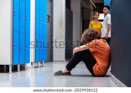 Sad caucasian teen boy sitting on floor in high school corridor while his classmates observe him. Copy space. Bullying consequences concept. Juvenile Depression Royalty-Free Stock Photo #2029938122