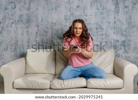 woman switching channels in front of TV at home. Stay home entertainments. Portrait  young  girl sitting on sofa, switches channels on the remote control and watching tv at home alone Royalty-Free Stock Photo #2029975241