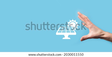 Hand on a blue background holds the icon symbol of solar panels. Renewable energy, solar panels station concept, green electricity.