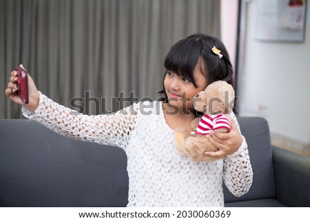 Cute little girl in white dress smiling as she takes a selfie of herself with smartphone. 10 years old child expressing happiness, embracing teddy bear for a picture. 