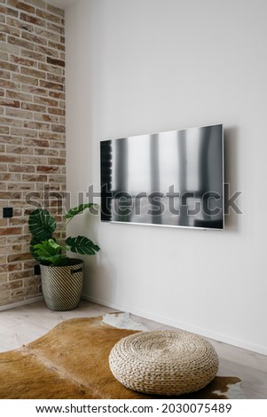 Vertical picture of big TV screen on clean white wall in modern living room with straw ottoman on cow skin rug and green potted plant in corner. Television in modern house or apartment