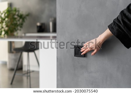 Cropped shot of woman turning light on or off in kitchen using black switch located on grey wall, modern kitchen interior design with plants, furniture and appliances in blurred background Royalty-Free Stock Photo #2030075540
