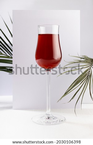 Red wine in elegant glass with summer tropical green palm leaf in sunlight with striped shadows in white abstract interior in minimal geometric style, vertical.