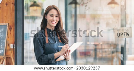 Beautiful asian young barista woman in apron holding clipboard and standing in front of the door of cafe with open sign board. Business owner startup concept.