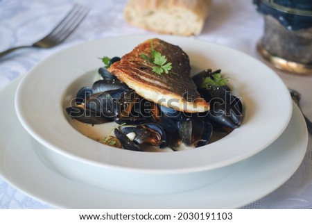 A pan sear crispy filet of sea bass fish on the sea mussels stew in white wine cream sauce in a white plate with parsley leaves on the top for a decoration  with fork and bread in background 