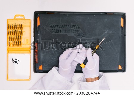 View from above of a white gloved hands of an engineer unscrewing the screws to open the lid of a laptop before being repaired on a white table