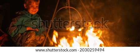 little four year old kid boy on a hike in the camp is watching the kettle is on the campfire in the night. bonfire with a burning flame in the dark and happy people. banner