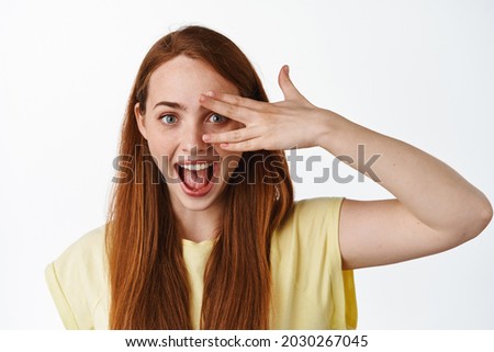 Close up portrait of attractive young ginger girl shows genuine happy emotion, peek through fingers and showing pure clean facial skin, white background