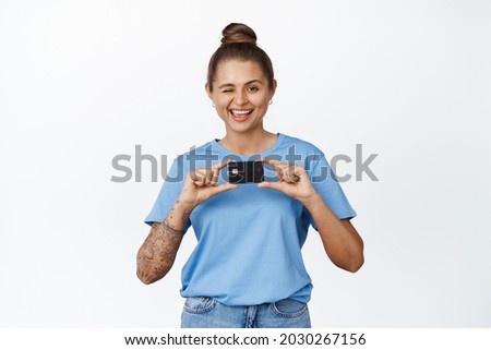 Happy young woman showing credit card, winking and smiling, standing over white background in blue t-shirt