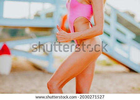 Beautiful perfect woman's body in pink bikini shining by body oil at the sand beach against lifeguard tower. Copy space.