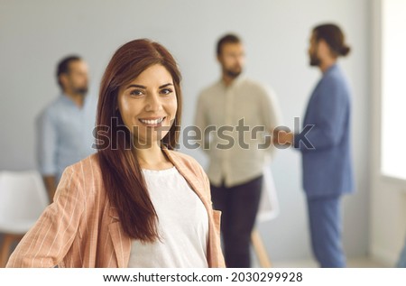 Close up portrait of a successful young business woman looking at camera with confident expression. Smiling woman on a background of people talking to each other in a bright room. Confidence concept.