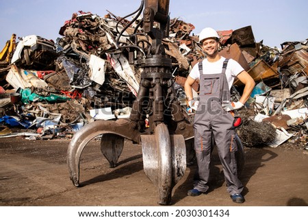 Portrait of worker standing by hydraulic industrial machine with claw attachment used for lifting scrap metal parts in junk yard. Royalty-Free Stock Photo #2030301434