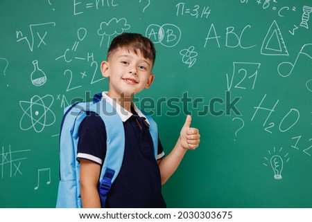 Young happy male kid school boy 5-6 years old in t-shirt backpack show thumb up gesture isolated on green wall chalk blackboard background studio. Childhood children kids education lifestyle concept.