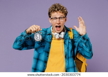 Young sad late boy teen student in casual clothes backpack headphones glasses hold clock isolated on plain pastel light violet background studio Education in high school university college concept.