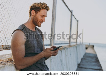 Young side view strong sporty athletic toned fit sportsman man 20s wear sports clothes warm up train use mobile cell phone at sunrise sun over sea beach outdoor on pier seaside in summer day morning.