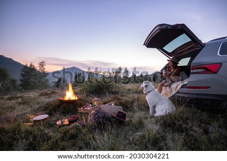 Woman enjoys beautiful view on the mountains, having a picnic with fireplace and sitting with dog in vehicle trunk at dusk on the evening. Traveling by car in nature Royalty-Free Stock Photo #2030304221
